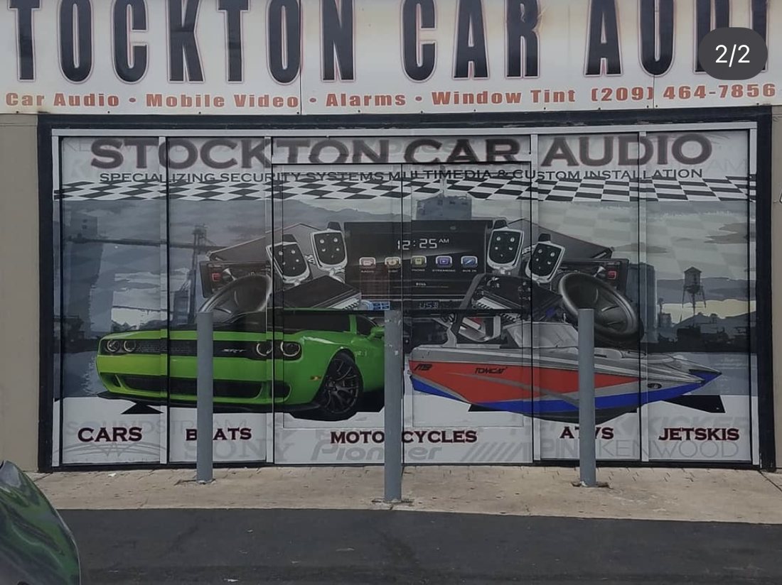 Stockton Car Audio storefront after: a lighter wrap across six glass panels with a green muscle car and a boat, head units and remotes, a Stockton waterfront silhouette, and the categories cars, boats, motorcycles, ATVs and jetskis along the base.