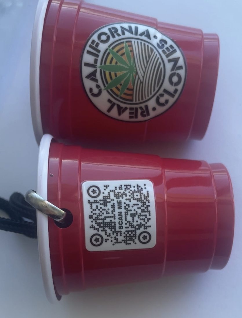 Two miniature red cups shown front and back: the roundel on one face, a scan-me QR code on the other, with a cord through a grommet.