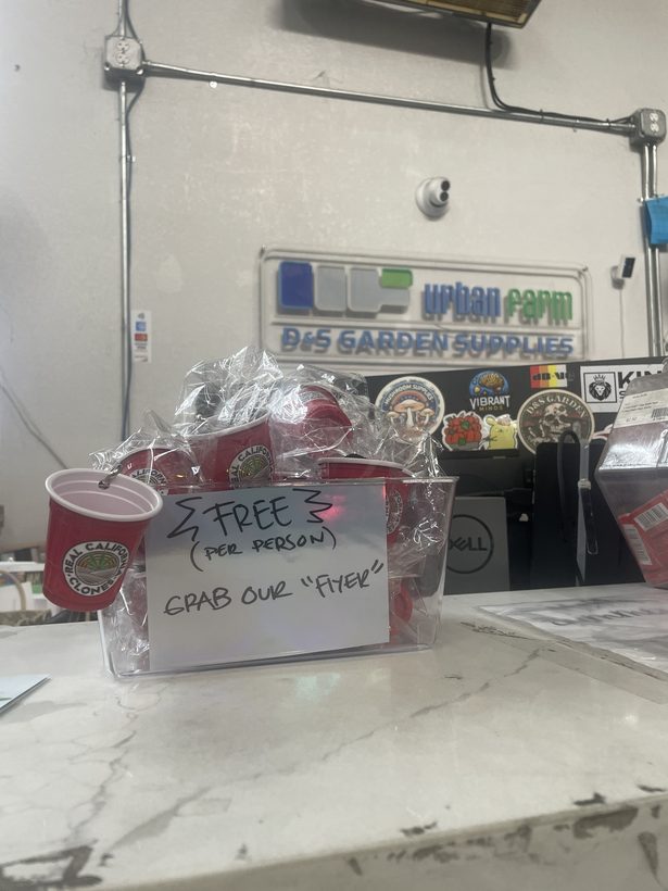 A clear bin of bagged mini cups on a garden supply shop counter with a handwritten sign reading free per person, grab our flyer.