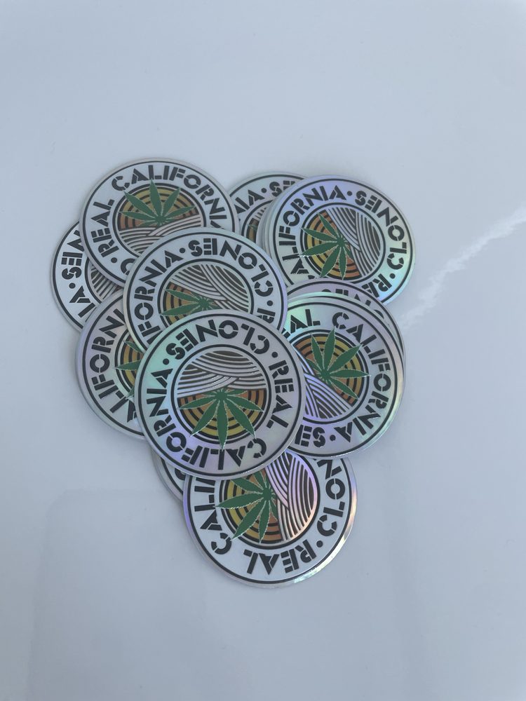 A stack of holographic Real California Clones roundel stickers catching rainbow light.
