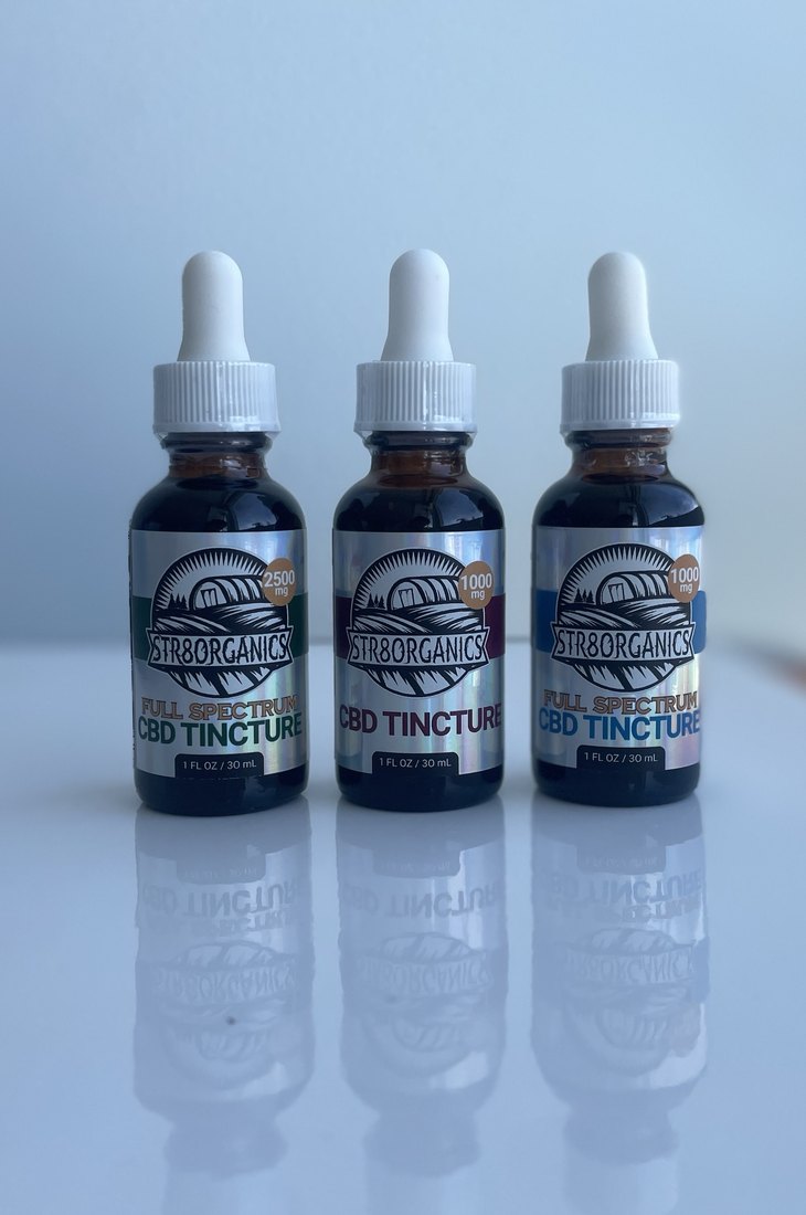 The three printed tinctures standing together on a reflective surface: 2500 mg full spectrum with a green band, 1000 mg no THC with a burgundy band, and 1000 mg full spectrum with a blue band, all on holographic labels.